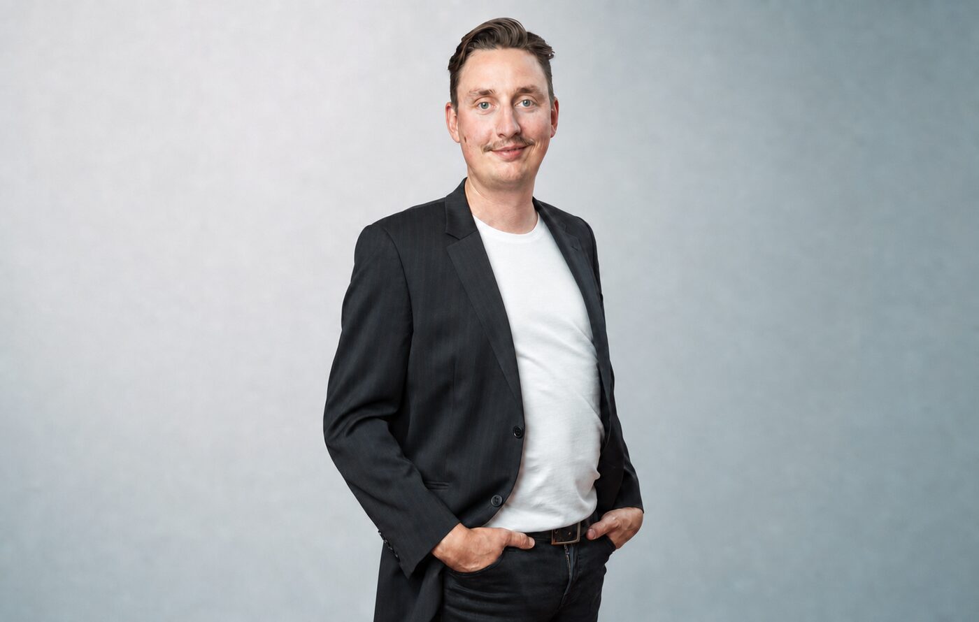 Jason Textor, Berater aus München, mit den Händen in den Hosentaschen vor einem grau-cyanfarbenen Hintergrund